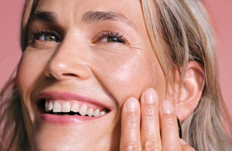 - Mulher sorridente de cabelos curtos grisalhos e pele hidratada passa niacinamida no rosto e perto das bochechas.