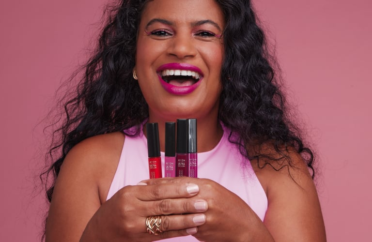 Uma mulher sorridente com batom rosa-choque e cabelos longos, escuros e ondulados segura três tubos de batom líquido em tons de vermelho e rosa da linha Power Stay da Avon. Ela veste uma blusa rosa-claro e posa contra um fundo rosa.