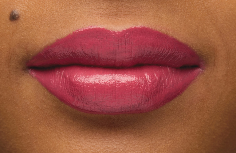 Close-up nos lábios de uma pessoa com batom rosa de acabamento cremoso. A pele apresenta uma pequena pinta acima do lábio superior.