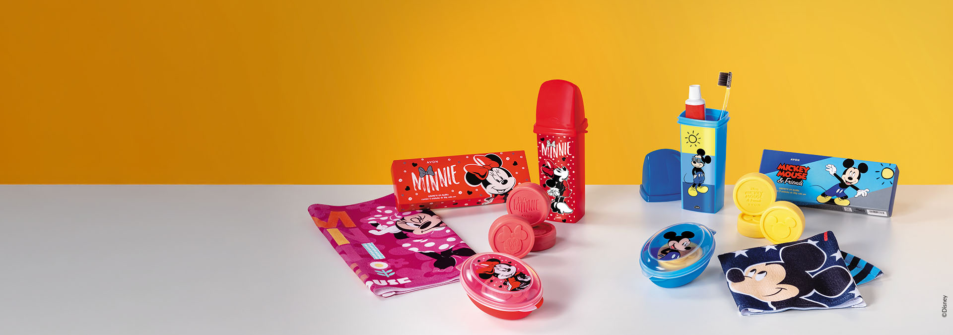 Banner em fundo amarelo. Em destaque, toalhas coloridas e itens de banho com estampas do Mickey e Minnie Mouse, como frascos e sabonetes. Texto: "Toalhas. Cores vibrantes que tornam o banho mais divertido."