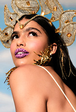 Mulher branca de cabelo preto comprido olhando sobre o ombro, em fundo de céu azul. Ela usa um adereço de cabeça dourado, delineado preto pontilhado e batom rosa com pontinhos escuros.