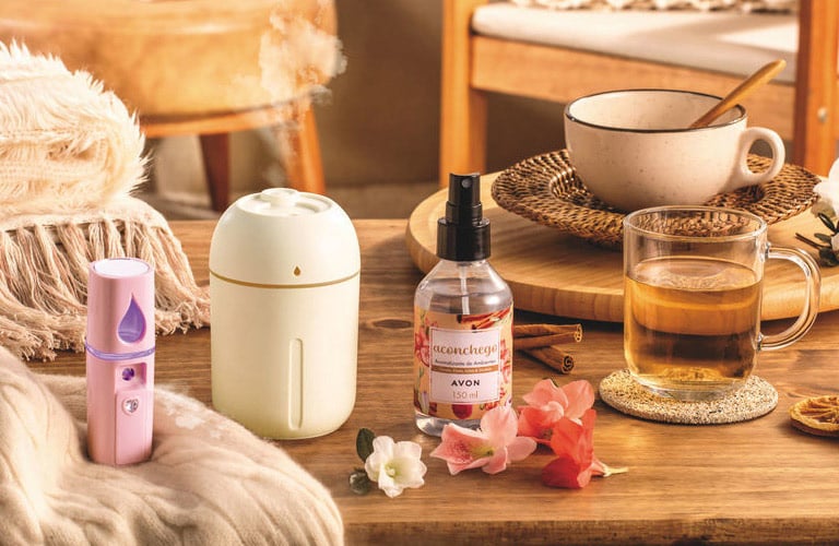 Uma mesa de madeira com manta rosa e difusor elétrico. Ao lado, um aromatizador de ambiente da Avon e poltrona com almofada estampada ao fundo.