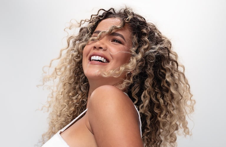 Mulher sorridente de cabelos cacheados com luzes loiras e esvoaçantes pelo vento, vestindo uma regata branca em um fundo neutro cinza.