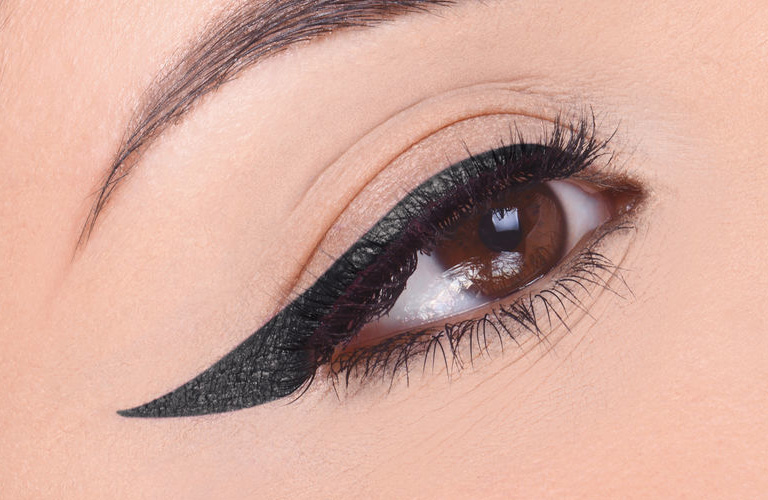 Close-up em olho castanho e amendoado com delineado preto gatinho e sombra rosa sutil na pálpebra. As bochechas têm sardas marrons claras.