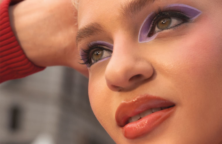 Mulher sorridente usa maquiagem colorida com sombra lavanda, batom terracota e blusa vermelha, posando com a mão no cabelo.