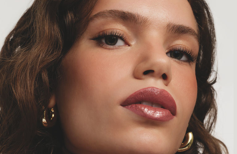 Mulher com cabelos ondulados castanhos, usa acessórios dourados e uma maquiagem marrom chocolate nos olhos e lábios.