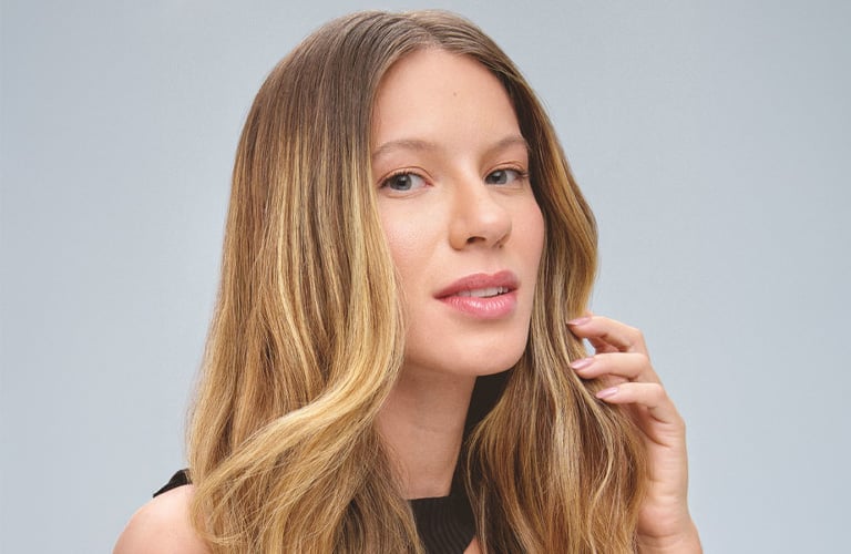 Mulher sorridente de cabelos loiros lisos e com leves ondas, vestindo uma regata preta contra um fundo cinza, enquanto toca suavemente as mechas de cabelo na altura do ombro com a mão esquerda.