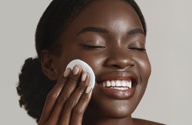 Mulher negra com a pele radiante, sorrindo com os olhos suaves enquanto passa um disco de algodão branco na bochecha. O fundo é neutro e em tom cinza-claro.