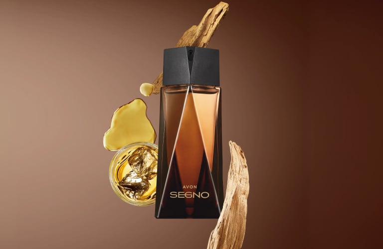 Perfume masculino, em tons de marrom e dourado com tampa preta, da linha Segno da Avon sobre um fundo marrom degradê. O frasco está cercado por pedaços de madeira e um copo com uma bebida alcoólica dourada e gelo.