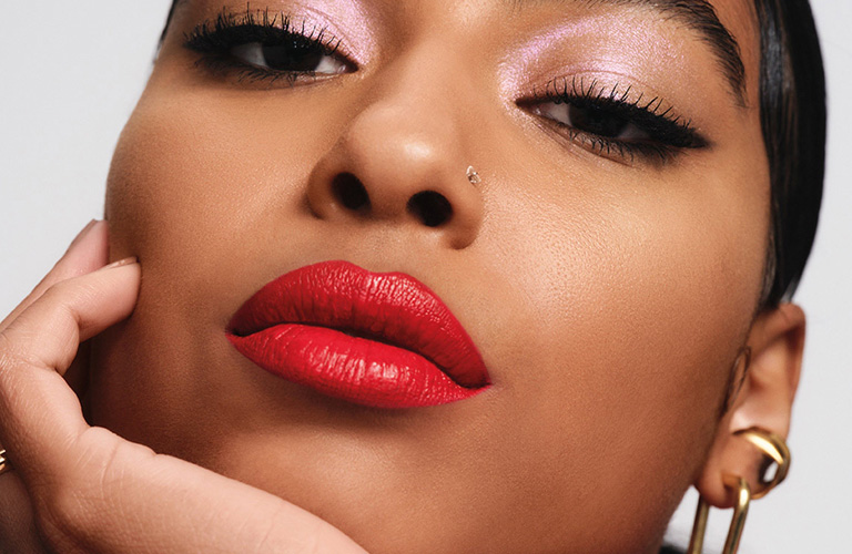 Mulher negra com batom vinil vermelho vibrante, sombra rosa cintilante e brincos dourados, posando com a mão no queixo.