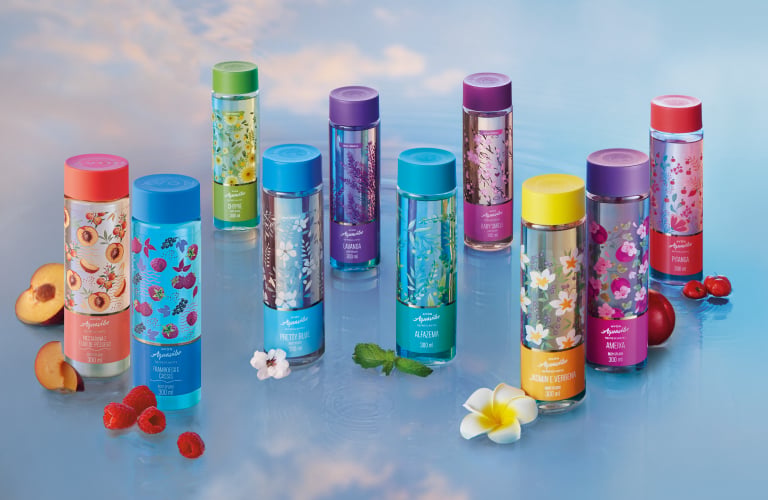 Frascos coloridos e estampados de body splashes da linha Aquavibe de Avon sobre uma superfície espelhada azul que reflete o céu ensolarado com nuvens ao fundo. Ao redor dos frascos, há pequenas frutas e flores decorativas.