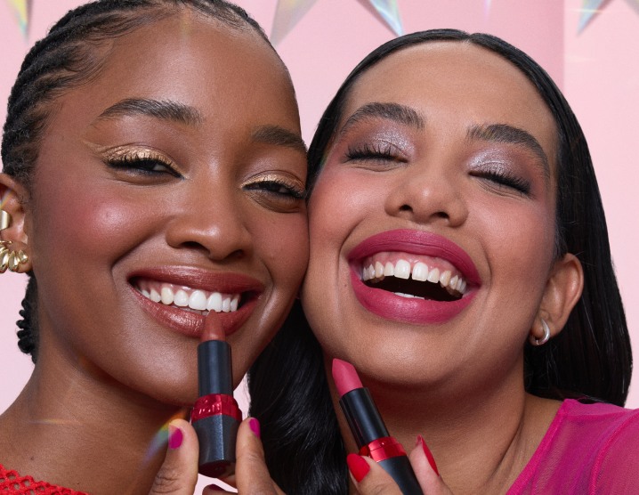 Mulheres com maquiagens iluminadas, sorrindo e segurando batons próximo aos lábios. À esquerda, mulher de cabelos trançados usando vermelho e, à direita, mulher de cabelos repartidos no meio usando rosa.