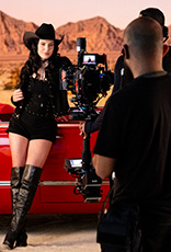 Mulher de cabelos longos castanhos, com roupa e chapéu pretos com pontos brilhantes, está à frente de carro conversível vermelho participando de gravação de vídeo. Fundo de montanhas desérticas com céu azul.