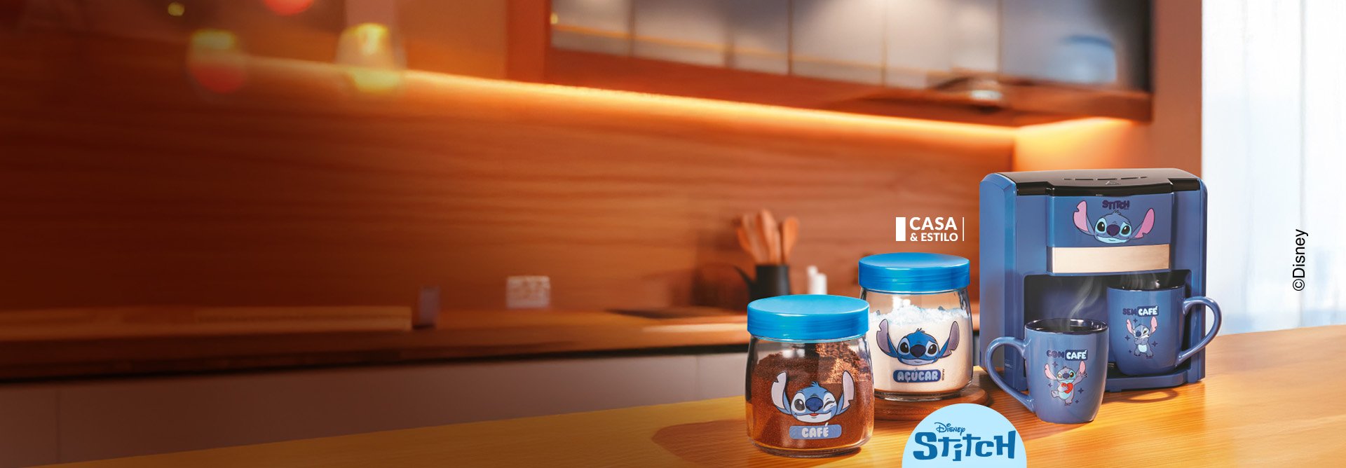 Itens Disney Stitch Casa & Estilo: dois potes de vidro para café e açúcar, máquina de café azul com duas canecas azuis sobre balcão de madeira, com fundo de madeira e iluminação quente. Texto ©Disney na lateral.