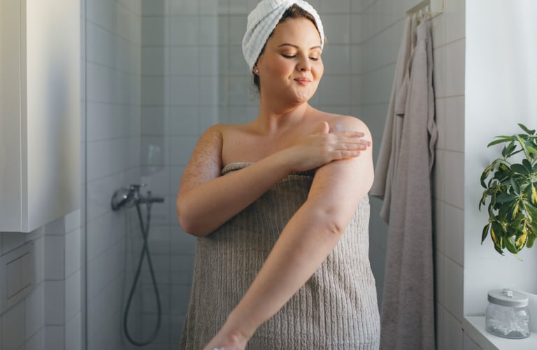 Mulher sorridente com uma toalha enrolada na cabeça e outra no corpo, aplicando hidratante corporal no braço após o banho e dentro de um banheiro iluminado.