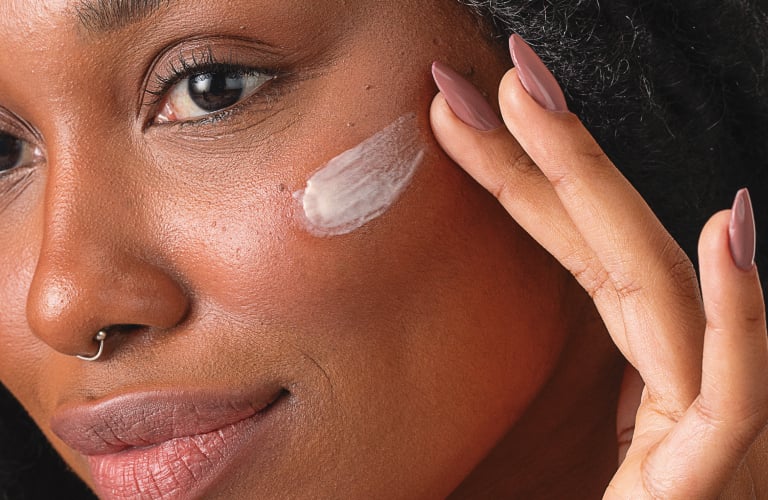 Mulher de pele negra com uma expressão suave e serena aplica com as pontas dos dedos uma camada de creme na bochecha, estendendo o movimento em direção à área abaixo dos olhos nas suas olheiras pigmentares. Ela tem cabelos longos presos em tranças escuras e usa um pequeno piercing no septo nasal.