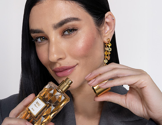 Mulher branca de cabelos lisos segura frasco da fragrância Topaze Elegant com tampa dourada e líquido pêssego. Ela usa roupa cinza e está em fundo claro.