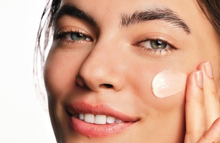 Mulher sorridente de cabelos e olhos castanhos usa um sérum de ácido hialurônico na região das bochechas.