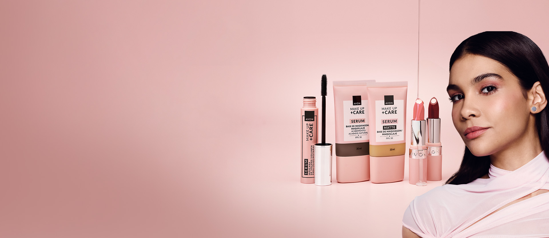 Em fundo rosa-claro, embalagens rosa Tratamake: máscara para cílios aberta, 2 bisnagas de tons de base e 2 batons, rosa e vinho, com textura incolor no batom rosa. À direita, mulher de cabelos castanhos.
