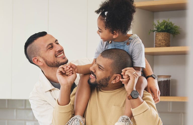Dois homens sorridentes de barbas e cabelos raspados se abraçam (um à esquerda e outro à direita). O homem à direita carrega uma criança sorridente nos ombros. As pessoas estão em uma cozinha com bancada branca, fruteiras e prateleiras de madeira.