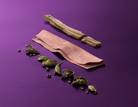 Amostras de cardamomo, couro e sândalo, ingredientes nobres da fragrância Black Essential Charm, aparecem em destaque no centro da tela sobre fundo violeta.