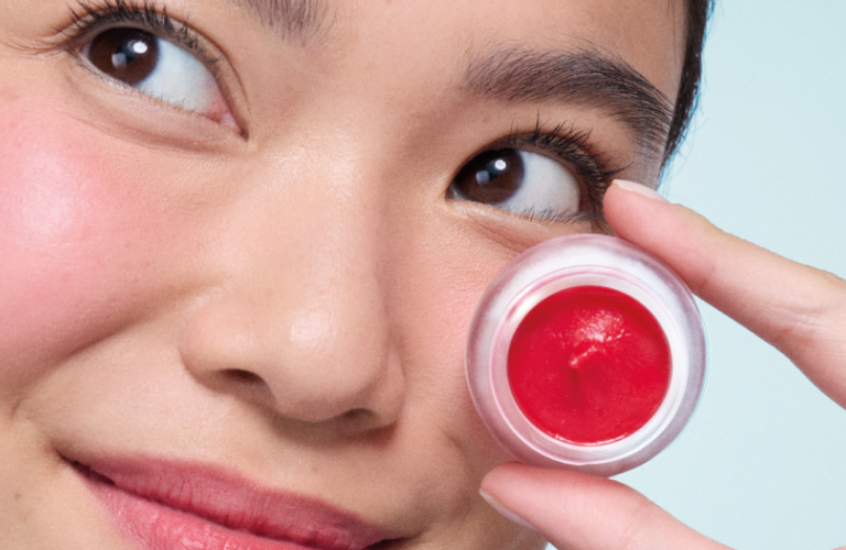 Mulher de bochechas e lábios intensamente rosados olha para cima e para o lado, segurando um pequeno pote transparente de balm labial em creme de cor vermelha vibrante, que está aberto e posicionado próximo ao seu olho.