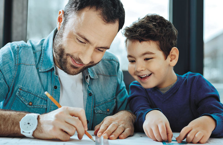 Um homem e um menino, provavelmente pai e filho, juntos em uma mesa de trabalho. O homem, vestindo jaqueta jeans e camiseta branca, segura um lápis e uma régua enquanto desenha em uma folha de papel. Ao seu lado, o menino, com um suéter azul, observa a atividade com um sorriso.