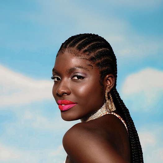 Mulher negra de cabelo trançado olha sobre o ombro, em um fundo de céu azul. Ela usa brinco dourado e batom matte vibrante, com aplicação meio a meio em tons de rosa e vermelho.