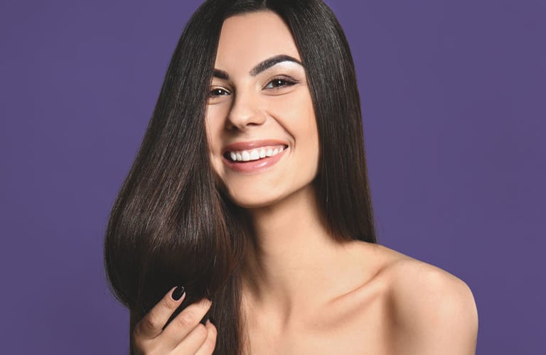 Mulher sorridente de cabelos castanhos lisos e longos, segura as pontas dos fios indicando a resistência do cabelo.