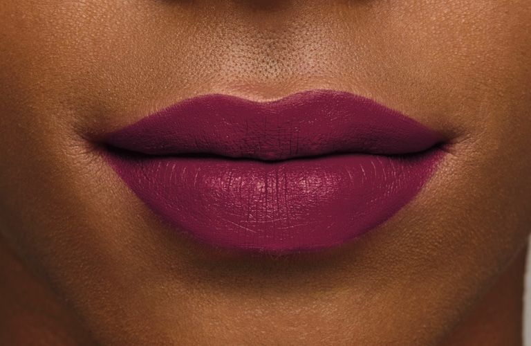 Close-up focado nos lábios de uma pessoa negra usando batom matte magenta fechado. A cor é uniforme e o acabamento é opaco.
