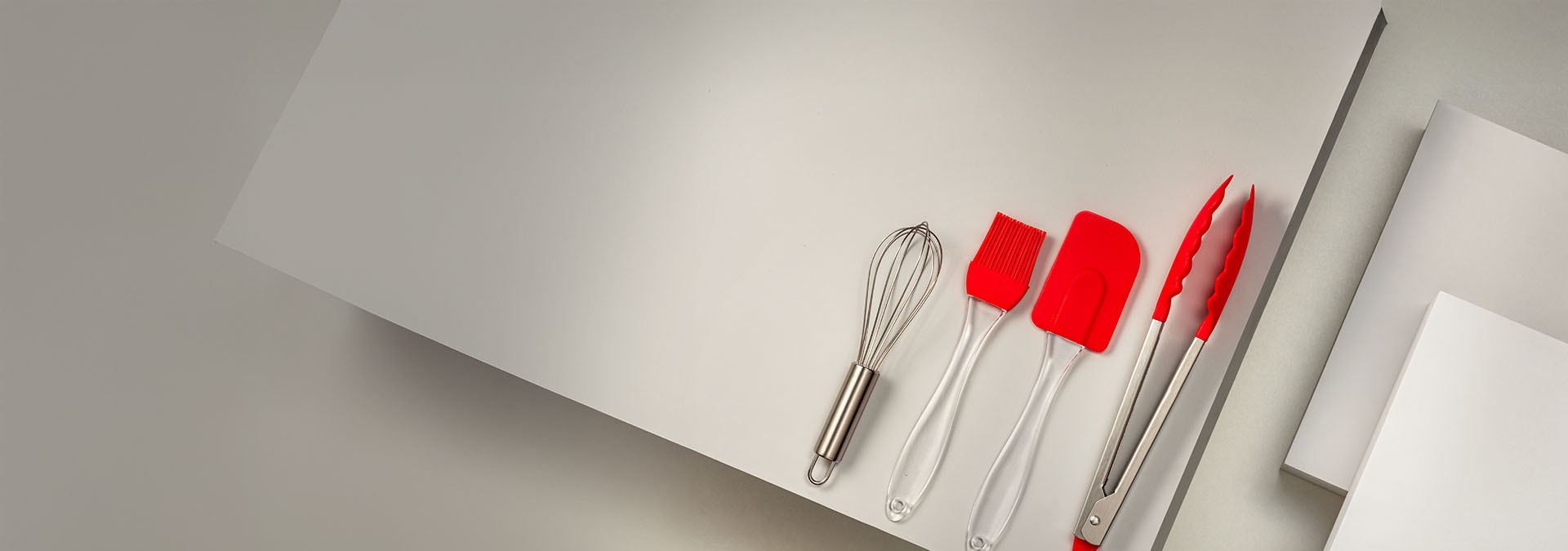 Banner em fundo claro. Em destaque,ílios de cozinha como um batedor, pincel, espátula e pegador, todos com detalhes em vermelho. Texto: "Utensílios. Itens práticos que tornam a cozinha mais divertida."