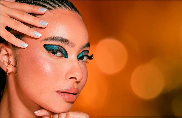 Mulher branca com maquiagem de carnaval com glitter. Os olhos tem sombra gráfica brilhante em tons de verde-esmeralda e azul-turquesa que envolve toda a pálpebra. Suas unhas com um esmalte prata metalizado, e o cabelo está preso em tranças.