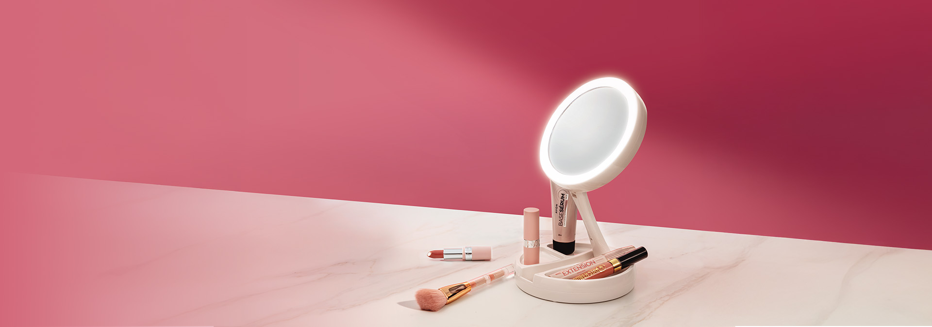 Banner com produtos de cuidados pessoais sobre fundo rosa. Inclui espelho iluminado, batons, base e pincel. Texto: "Cuide-se com carinho! Descubra nossos produtos que realçam sua beleza."