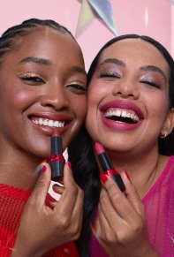 Mulheres com maquiagens iluminadas, sorrindo e segurando batons próximo aos lábios. À esquerda, mulher de cabelos trançados usando vermelho e, à direita, mulher de cabelos repartidos no meio usando rosa.