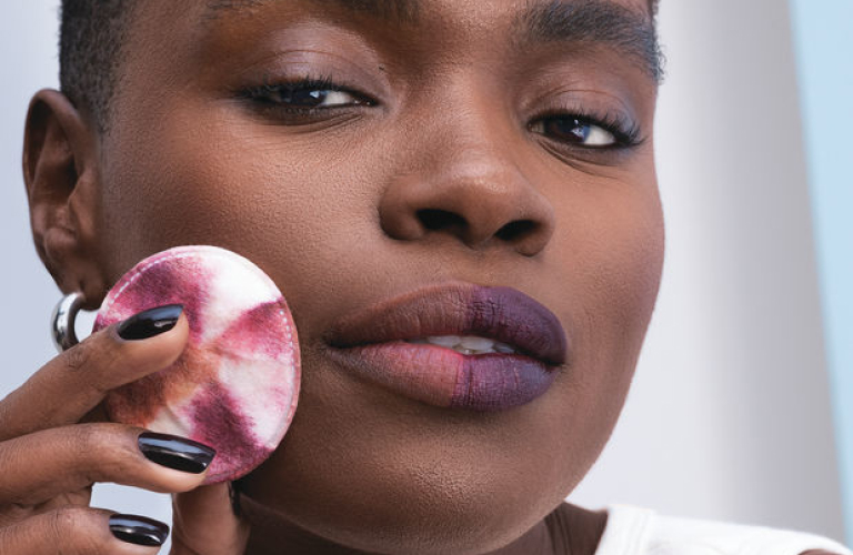 Mulher negra usa água micelar em um algodão para retirar a maquiagem dos lábios, olhos e rosto, Ela segura o algodão sujo em frente ao rosto.