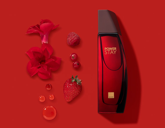Frasco de fragrância Power Stay em vermelho sobre fundo vermelho com ingredientes como flores, framboesa,  mirtilo e morango, à esquerda.
