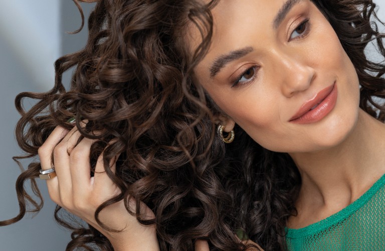 Mulher branca com cabelos cacheados castanhos de curvatura 3C e a cabeça levemente inclinada para esquerda, apertando com as mãos as pontas dos fios para finalizar o cabelo.