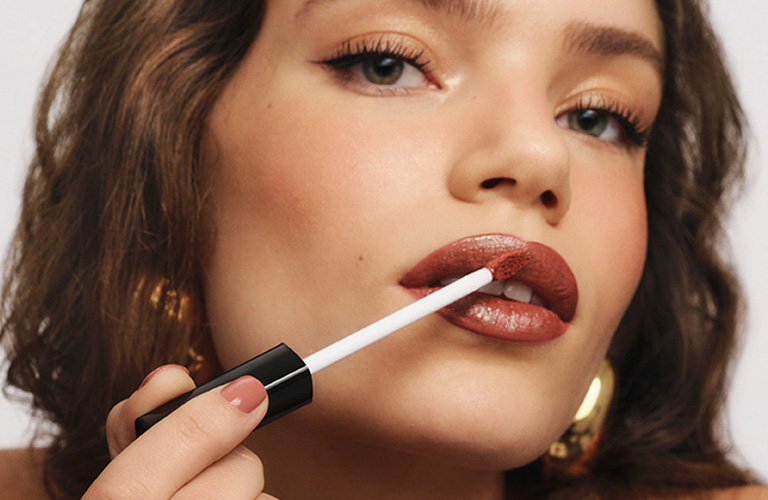 Mulher jovem passando batom marrom acobreado com um aplicador. Ela tem cabelos ondulados, usa brincos dourados e uma maquiagem leve com delineado fino.