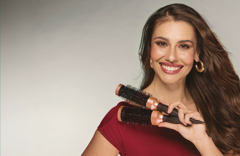 Mulher sorridente de cabelos castanhos ondulados jogados para o lado esquerdo, vestindo uma blusa de manga curta vinho e usando brincos de argola dourados. Com as mãos cruzadas à frente do corpo, ela segura duas escovas de cabelo modeladoras redondas, pretas com detalhes dourados.
