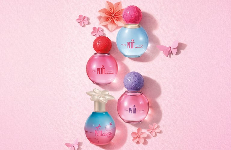 Quatro perfumes para adolescentes da linha Petit da Avon. Os frascos têm cores azul, rosa e degradê, e são decorados com tampas esféricas coloridas e uma tampa em formato de flor transparente. Eles estão organizados sobre uma superfície rosa-claro texturizada e cercados por pequenas flores.