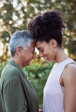 Mulheres negras de idades diferentes, frente a frente com as testas encostadas. À esquerda, mulher de cabelos curtos grisalhos de verde e, à direita, mulher de cabelos presos e crespos de rosa. Fundo externo doméstico.