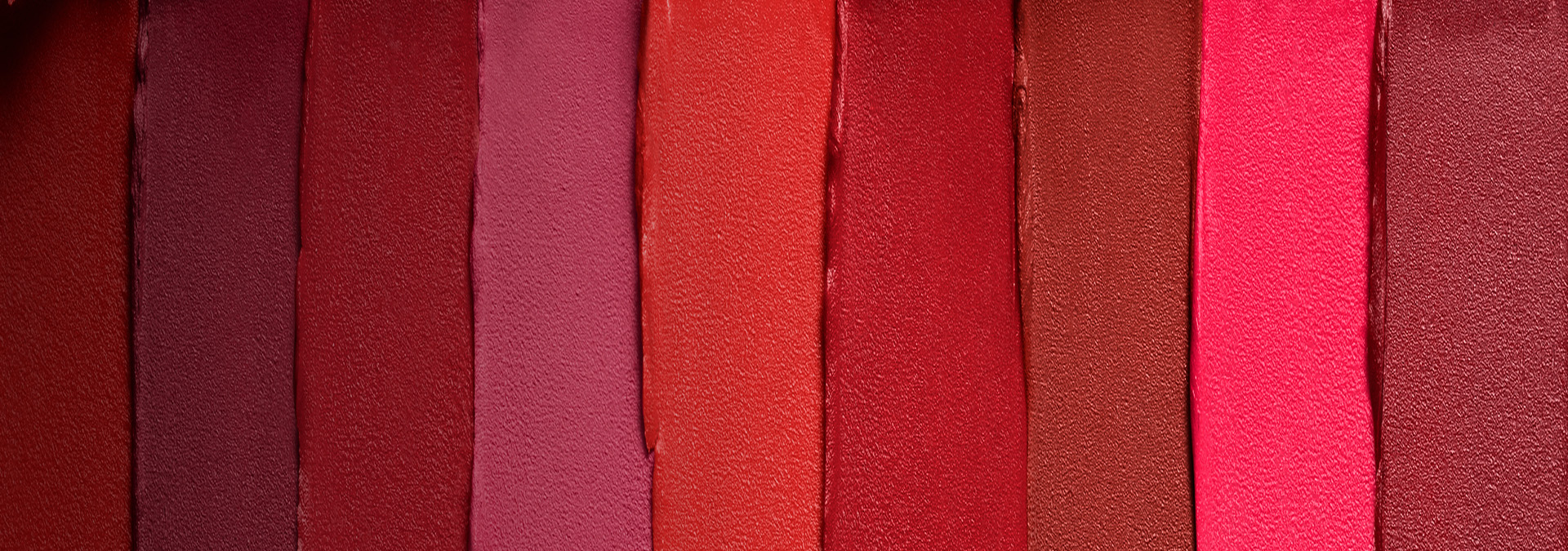 Banner com fundo em tons de vermelho e rosa. Elementos incluem faixas de batons aplicados em diferentes tonalidades, formando um degradê vibrante. Texto: "Maquiagem: mais presentes, mais emoção."