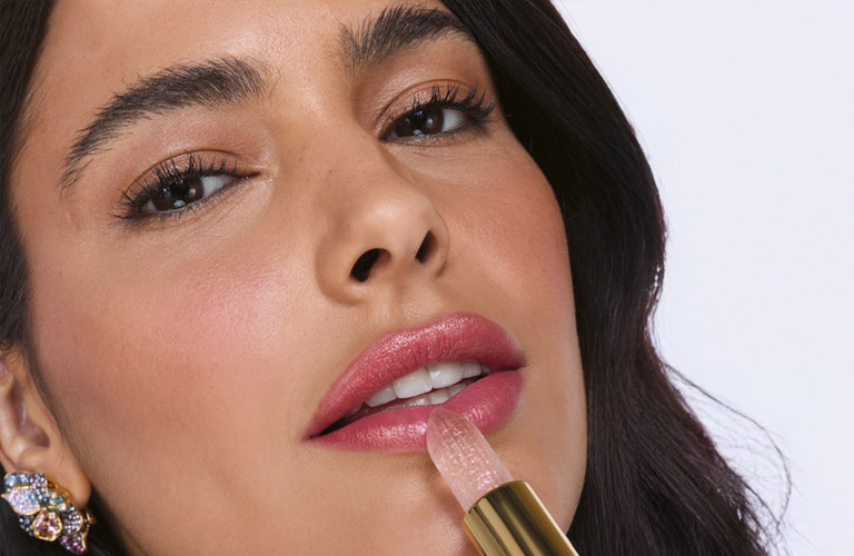 Close-up de mulher de cabelos castanhos e brincos de pedrarias, passando um batom rosa claro que reage ao pH dos lábios. Ela usa uma maquiagem de noiva simples em tons rosados.