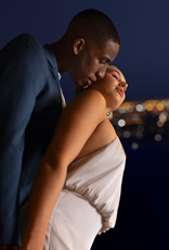 Casal apaixonado em fundo com o horizonte de uma cidade iluminada. Ela está com vestido branco e de costas para ele que está de terno azul.