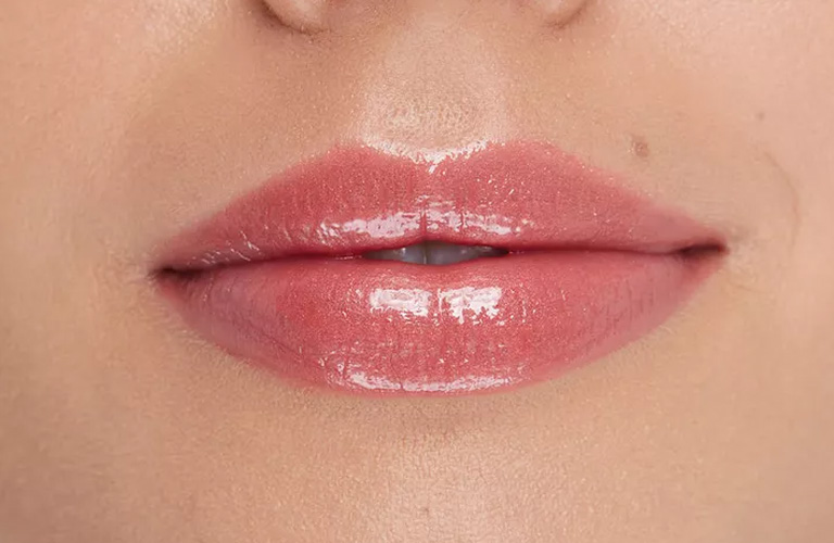 Close-up nos lábios de uma pessoa usando gloss labial rosado e translúcido. O acabamento é brilhante e molhado e os lábios estão fechados.