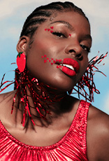 Mulher negra de cabelo trançado, usando batom vermelho, brincos de morango e blusa vermelha brilhante. Sua maquiagem em tons de vermelho traz corações nos olhos e ao redor dos lábios. Fundo azul.