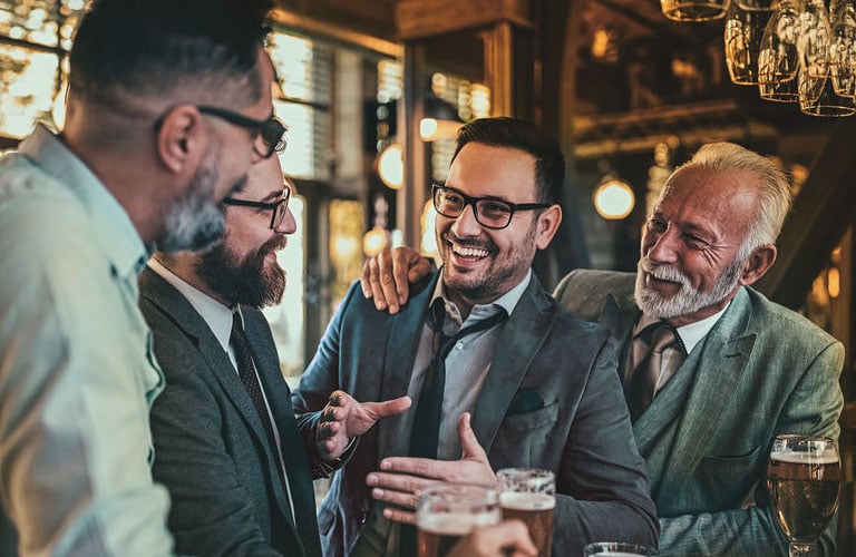 Quatro homens de diferentes idades estão sorridentes e cumprimentam-se em um bar, vestindo roupas formais. Cada um tem um estilo de barba (curta, cheia ou cavanhaque).