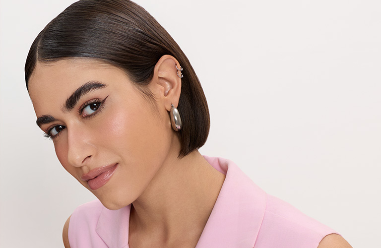 Mulher negra sorridente com batom avermelhado de brilho suave. Ela usa argolas prateadas com detalhes coloridos e uma camiseta azul.