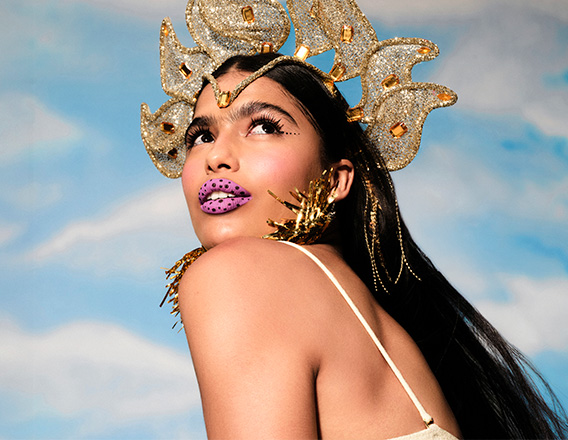 Mulher branca de cabelo preto comprido olhando sobre o ombro, em fundo de céu azul. Ela usa um adereço de cabeça dourado, delineado preto pontilhado e batom rosa com pontinhos escuros.