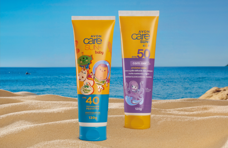 Dois tubos de protetor solar infantil da linha Avon Care Sun posicionados sobre a areia da praia, com o mar azul e o céu ensolarado ao fundo. O tubo em primeiro plano é amarelo e azul, enquanto o tubo ao fundo é amarelo e roxo.